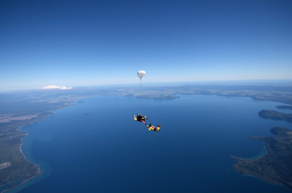 Two skydivers freefall above the vast, deep blue expanse of Lake Taupō in New Zealand, with a small white parachute deploying against a clear sky and distant horizon.