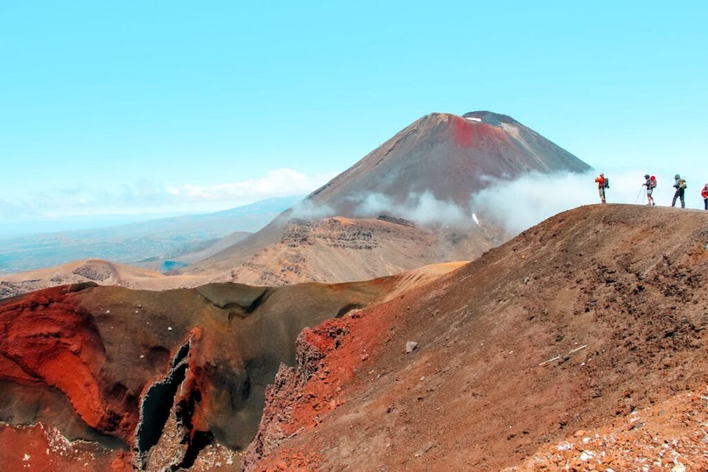 Hikers traverse the rugged volcanic landscape of the Tongariro Alpine Crossing in New Zealand, with Mount Ngauruhoe (Mount Doom) rising in the background under a bright blue sky.
