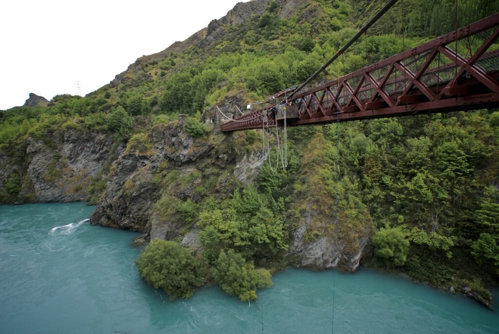 A scenic view of the historic Kawarau Bridge in Queenstown, New Zealand, with bungee cords hanging over the turquoise river below, surrounded by rugged cliffs and lush greenery.