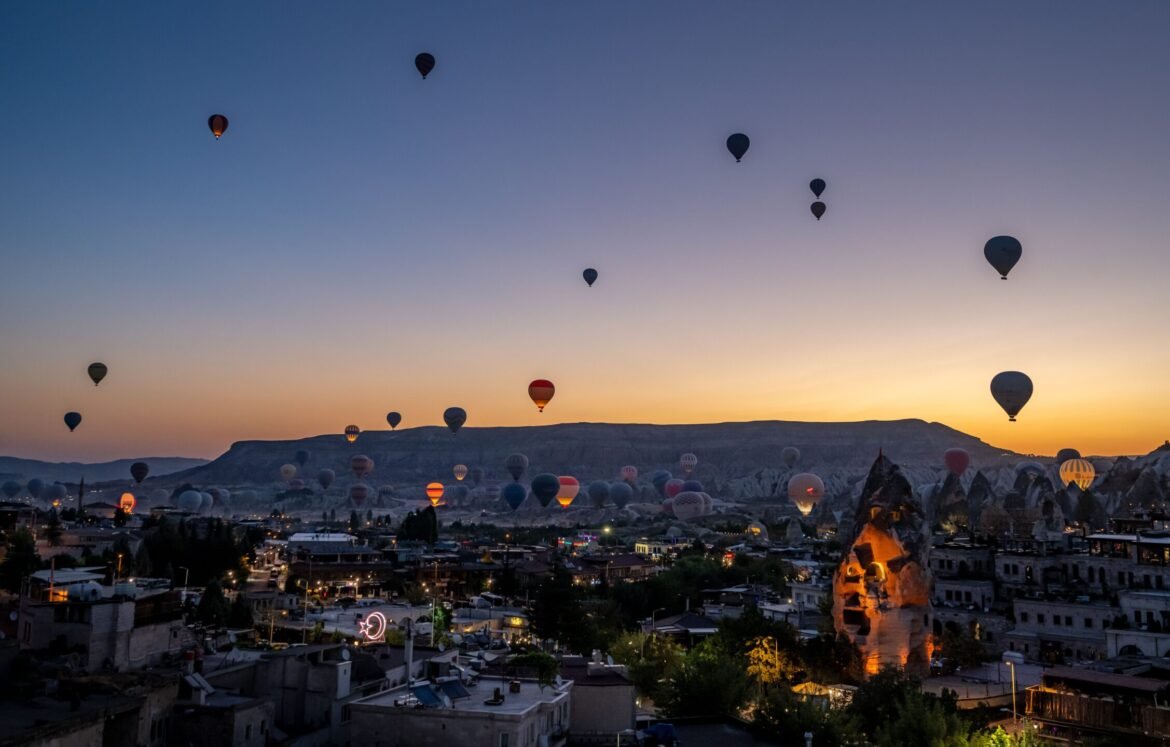 A stunning view of hot air balloons rising over the city of Cappadocia at sunrise, with the unique rock formations and landscape in the background.