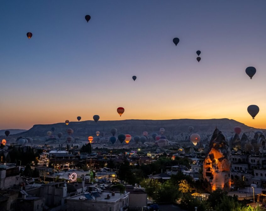 A stunning view of hot air balloons rising over the city of Cappadocia at sunrise, with the unique rock formations and landscape in the background.