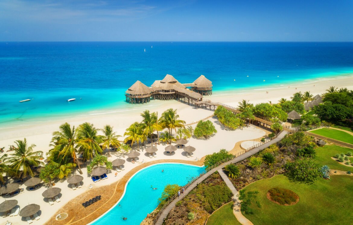 Aerial view of beautiful hotel on the sea, pool, umbrellas and green palm trees on the sandy beach at sunny summer day. Top view. Tropical landscape with wooden hotel on the blue water, trees, boats