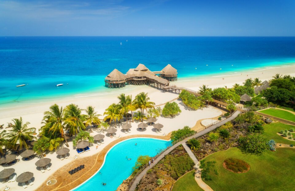 Aerial view of beautiful hotel on the sea, pool, umbrellas and green palm trees on the sandy beach at sunny summer day. Top view. Tropical landscape with wooden hotel on the blue water, trees, boats