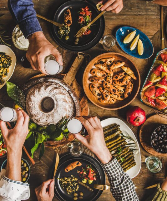 Traditional Turkish celebration dinner. Flat-lay of people eating Turkish salads, cooked vegetables, meze starters, pastries and drinking raki drink, top view, wide composition. Middle Eastern cuisine