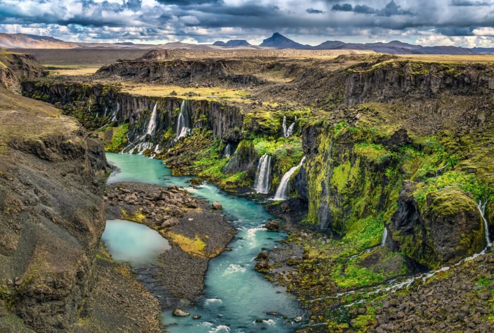 Scenic view of a dramatic Icelandic canyon with waterfalls cascading into a turquoise river under a cloudy sky.